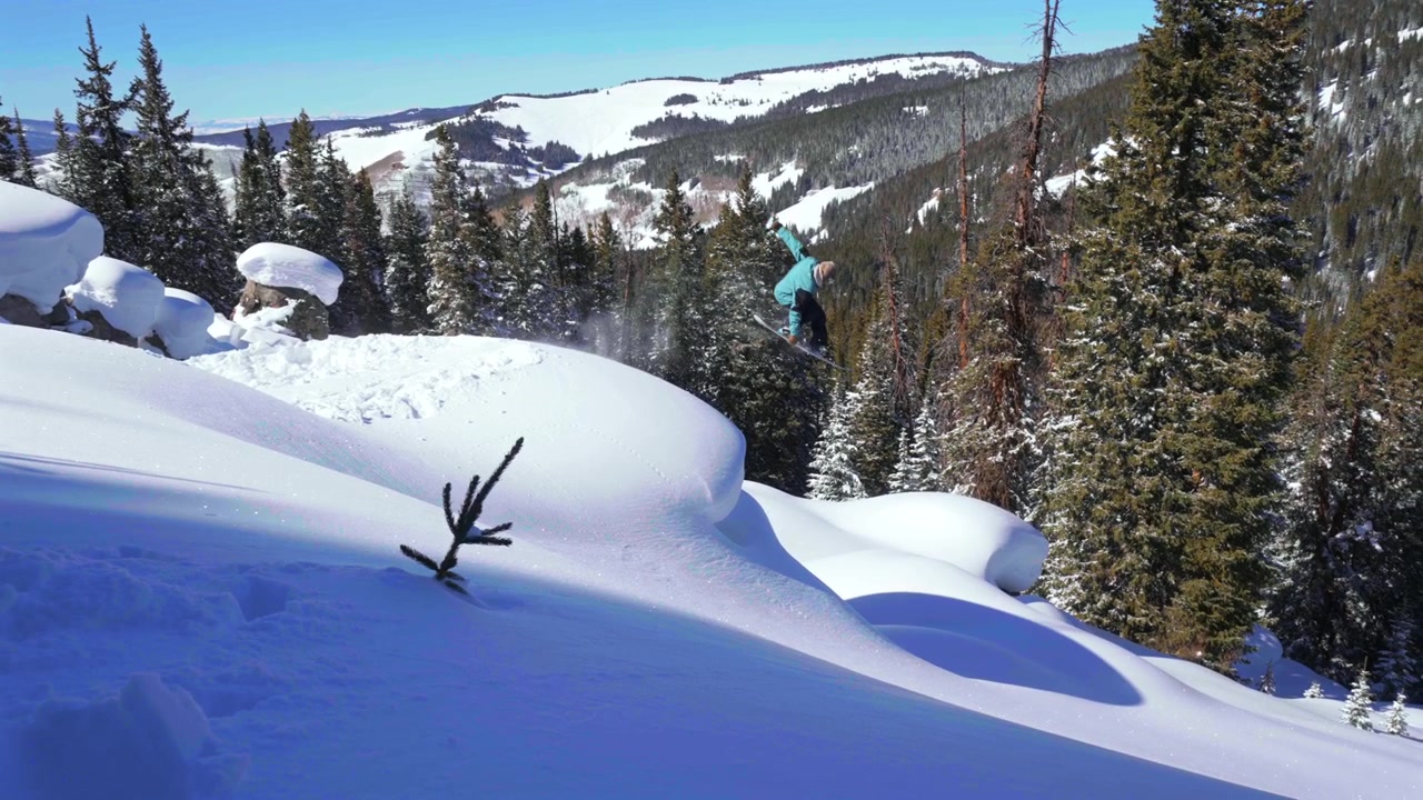 Snowboarder professionista che pratica snowboard fuoripista su cuscini di neve fresca e dirupi, in inverno a Vail Pass, Colorado, scendendo le ripide Montagne Rocciose. Neve fresca e profonda, salto, Indy grab, trick aerei, cielo blu, mattina soleggiata, con effetto panning.