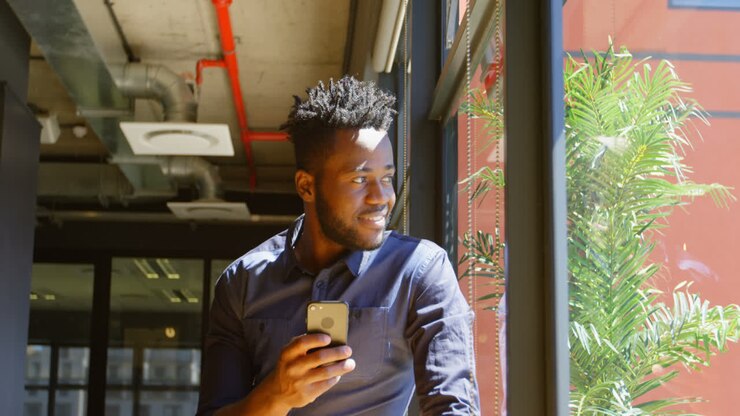 Front view of happy young black businessman looking through window while using mobile phone 4k