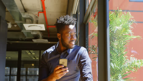 Vista-Frontal-De-Un-Joven-Y-Feliz-Hombre-De-Negocios-Negro-Mirando-Por-La-Ventana-Mientras-Usa-El-Teléfono-Móvil-4k