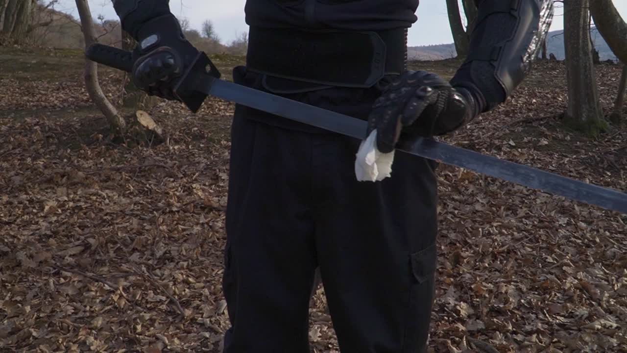 A stealthy warrior in black tactical gear carefully cleans his katana blade with a cloth. The setting is a forest covered in dry leaves, enhancing the cinematic and mysterious atmosphere