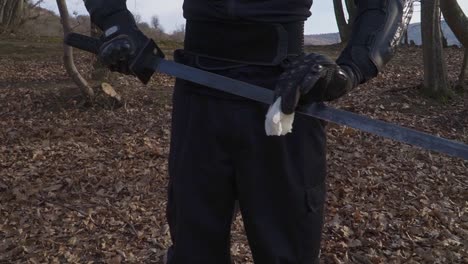 a stealthy warrior in black tactical gear carefully cleans his katana blade with a cloth. the setting is a forest covered in dry leaves, enhancing the cinematic and mysterious atmosphere