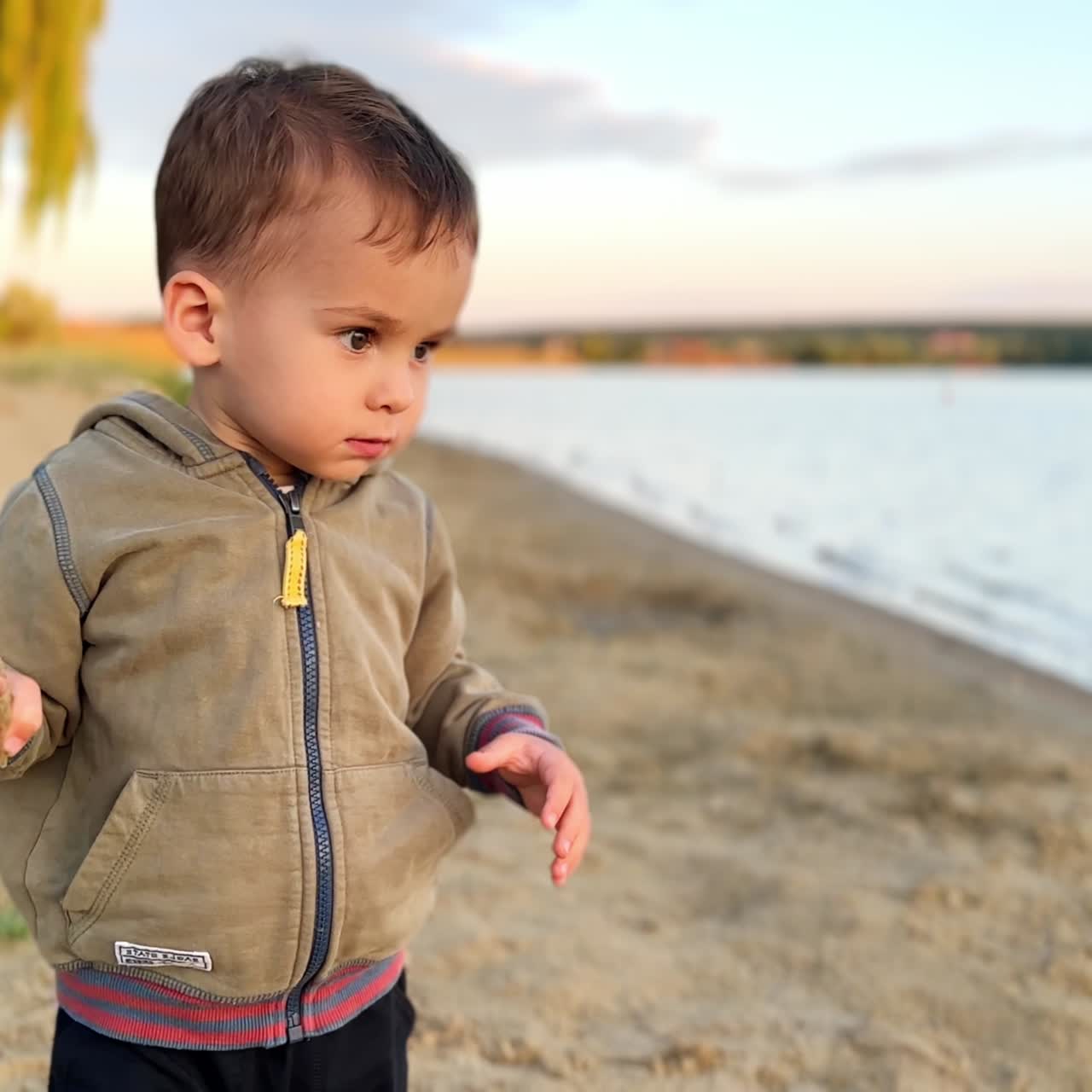 Cute Caucasian toddler on the river bank. Baby boy throws sand from the shore into water
