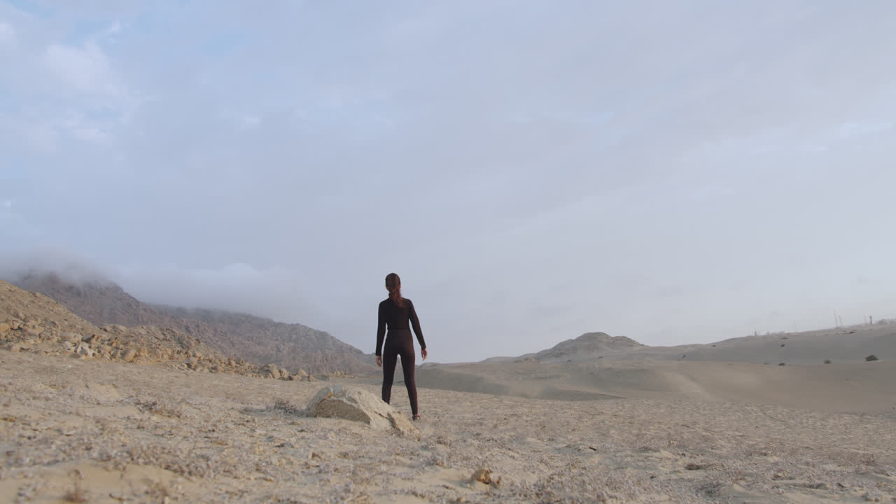 Campo lungo di una bella donna in nero in piedi accanto a una roccia nel mezzo del deserto alla luce del giorno.