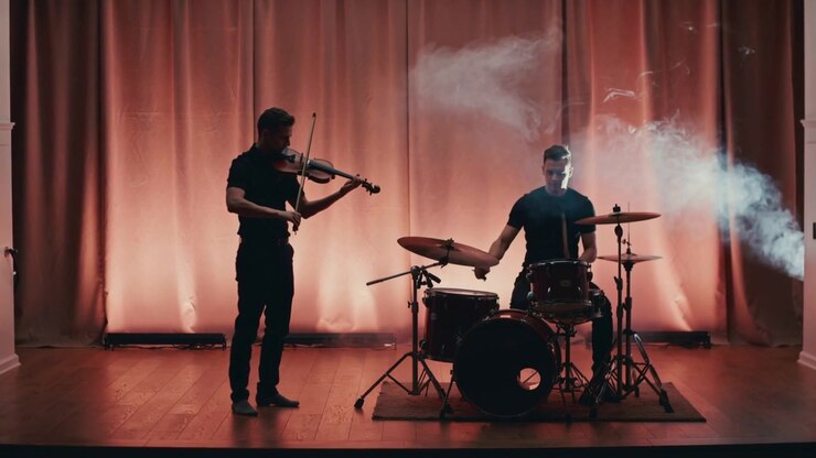 Violinista e batterista che suonano, il violinista che tira l'archetto e il batterista che colpisce rullante e piatti sul palco, con fumo che appare.