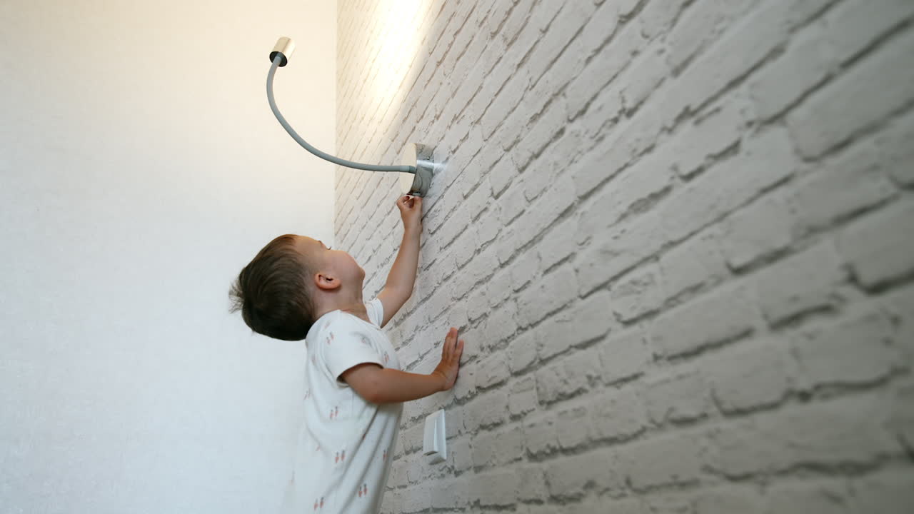 Cute baby boy stands near the white break wall holding for it. Toddler reaches the lamp above him to switch off the light. Low angle view.
