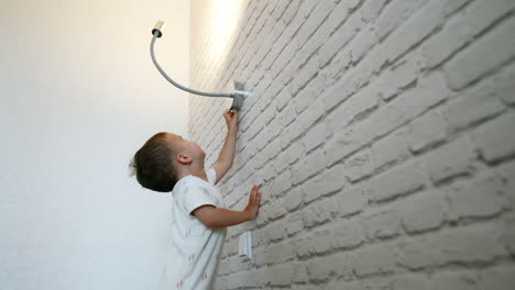 cute baby boy stands near the white break wall holding for it. toddler reaches the lamp above him to switch off the light. low angle view.