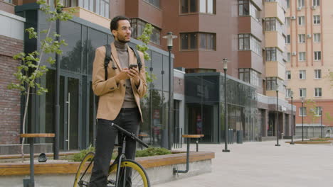 hombre americano sonriente con ropa formal usando teléfono móvil mientras está de pie con su bicicleta en la calle