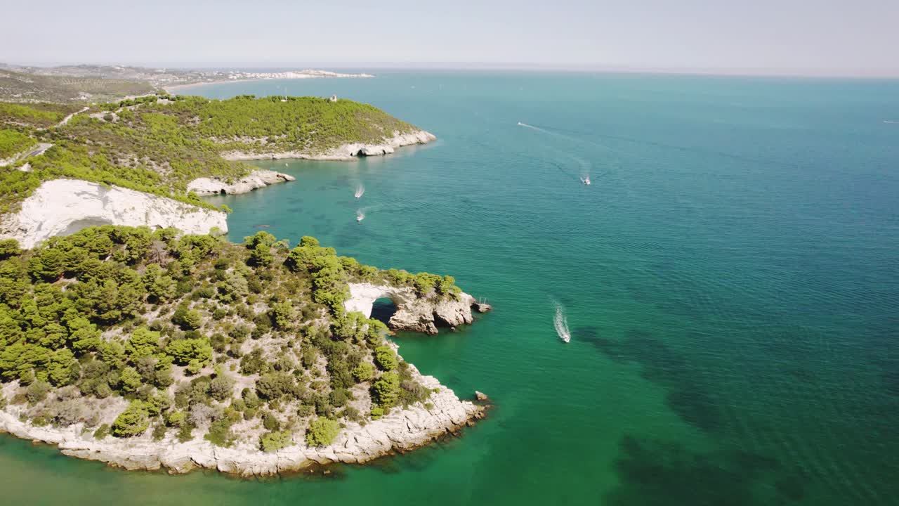 가르가노 국립공원의 해안선 위로 위쪽으로 날아가는 드론과 4k로 지나가는 보트와 바다의 아치를 보여줍니다.