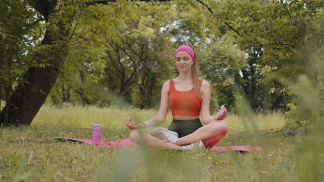 young woman sitting on mat in lotus position, relaxing, practicing yoga meditation in city park