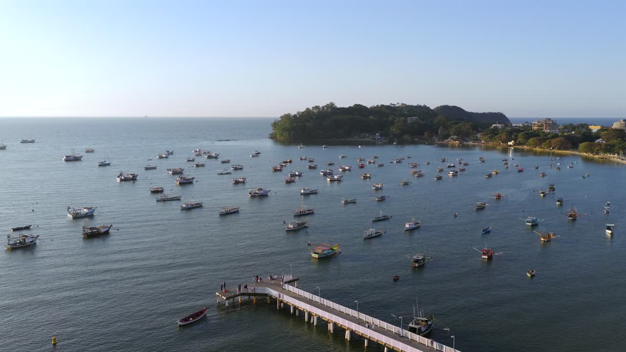 Praia do Trapiche in Penha, Santa Catarina, Brazil, with dozens of fishing boats anchored across the calm sea near the wooden pier at sunset, static drone view