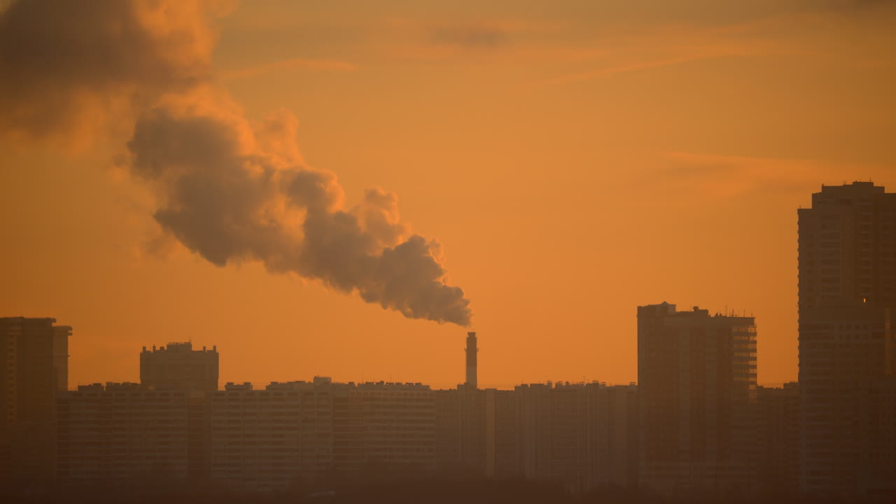 Industrial chimney releasing thick emissions above residential city skyline in warm orange sunset light Urban air pollution concept with dense haze high rise buildings and dramatic atmosphere