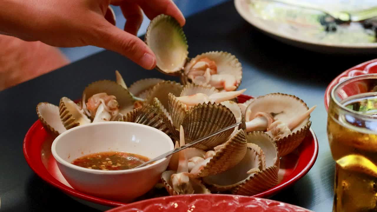 Hands open clams over a plate, dipping them in sauce. Warm lighting enhances the casual dining atmosphere