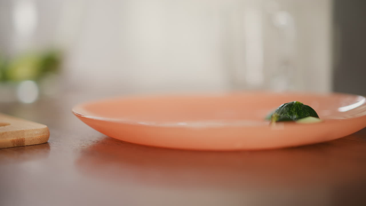 Close up of cucumber on orange plate placed on kitchen table as person picks full cucumber while leaving cut piece, soft focus background enhances natural kitchen ambiance