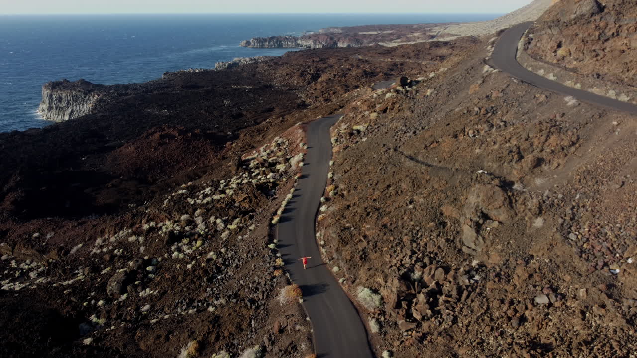 camino sinuoso alrededor de una alta montaña en las islas canarias, que corre a una mujer joven en un vestido, en los acantilados de fondo con el mar en una relajante puesta de sol, isla de hierro - tiro de drones