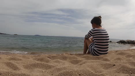 Scenic-cute-young-woman-sitting-on-beach-in-Sithonia-,-Greece,-static-rear