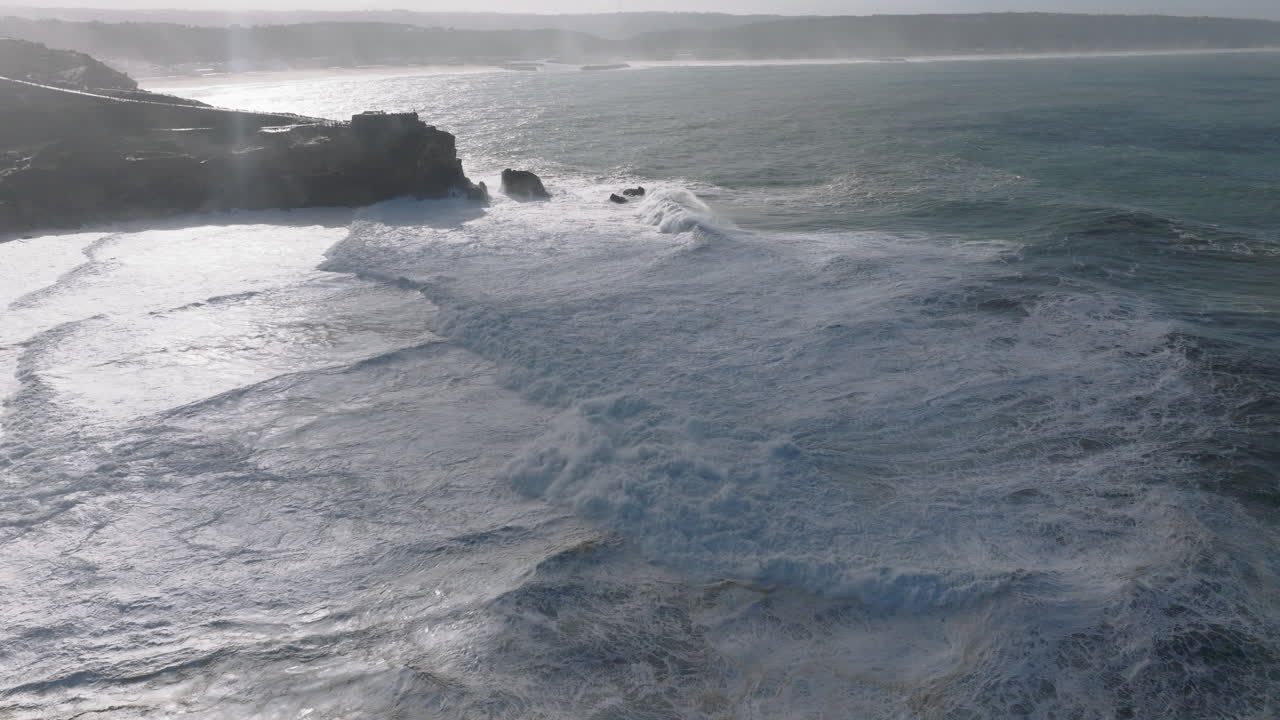 Aerial drone shot of waves coming into shore on a day with giant waves in Nazaré, Portugal, Europe. Farol da Nazaré lighthouse visible. Big wave surfing town and home of tow-in surfing