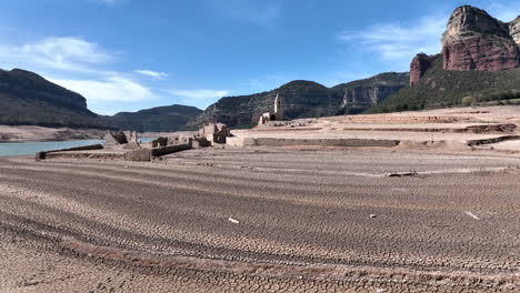 Aerial-flying-above-dry-cracked-soil-in-Sau-water-reservoir,-resurfaced-church