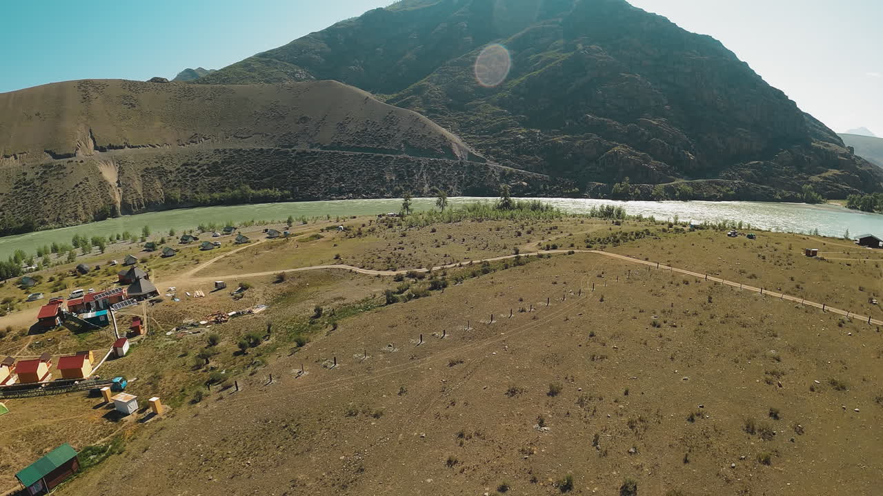 Majestic waters flow between undulating terrain. Serene presence of campsite along bank under benevolent gaze of clear sunny sky. River currents