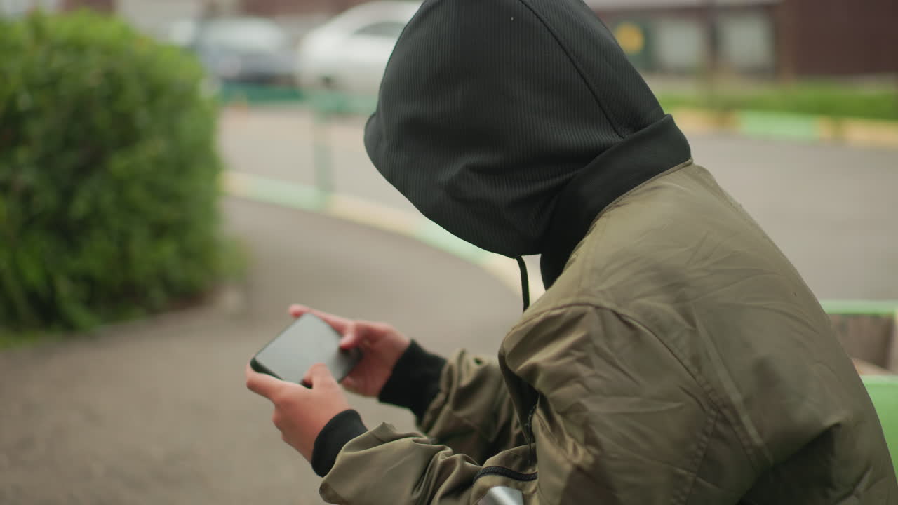 Rear angle view of boy in hooded jacket seated outdoors holding handheld device, focused on playing game, blurred greenery and parked cars in background giving casual urban feel