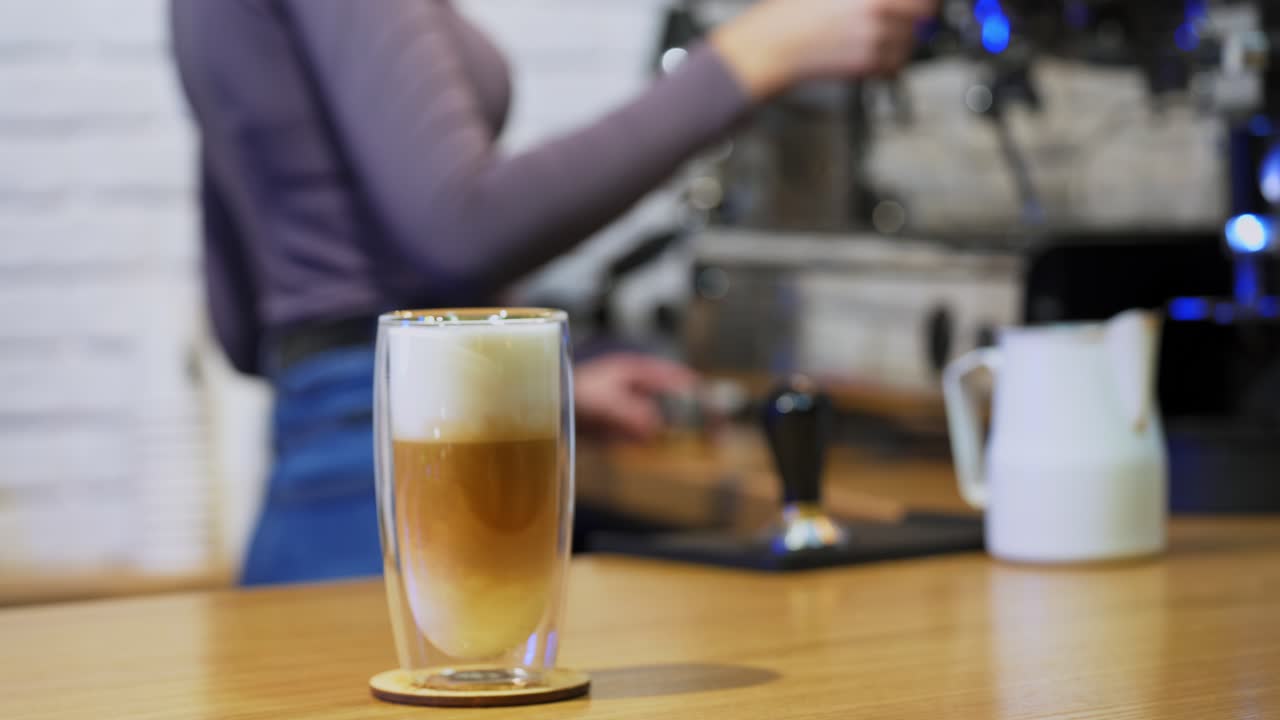 Cappuccino or latte in glass. Woman puts drink from coffee machine. Fresh coffee with whipped cream in tall clear glass on table. Close-up.
