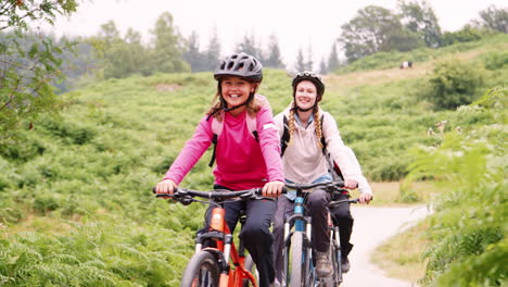 Young-family-having-fun-riding-mountain-bikes-on-a-country-lane-during-a-camping-holiday,-Lake-District,-UK