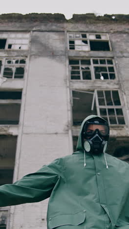 abandoned place and a person in safety suit. survivor in gas mask and hazmat suit standing with stretched arms on ruined building background. chemical catastrophe. vertical video