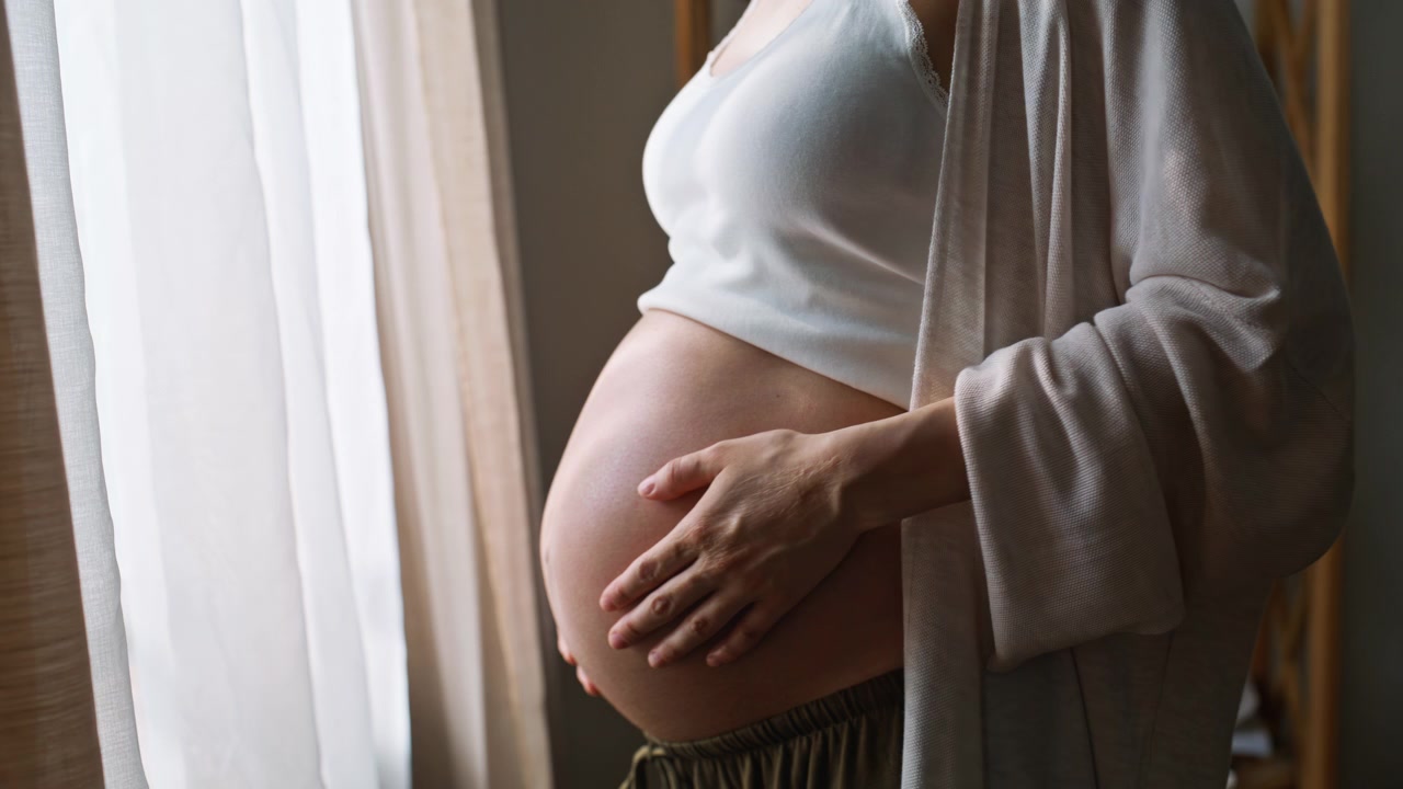 Futura mamma che si tocca i capelli vicino alla finestra. Primo piano del pancione tra le mani.