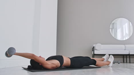 Beautiful-brunette-girl-lifts-her-torso-with-a-dumbbell-in-her-hands-to-her-leg-lying-on-a-rug-in-a-white-apartment.