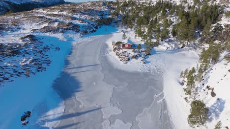 Fly-Away-Over-Wooden-Cabin-By-The-Frozen-River-Mountains-Near-Bessaker,-Norway