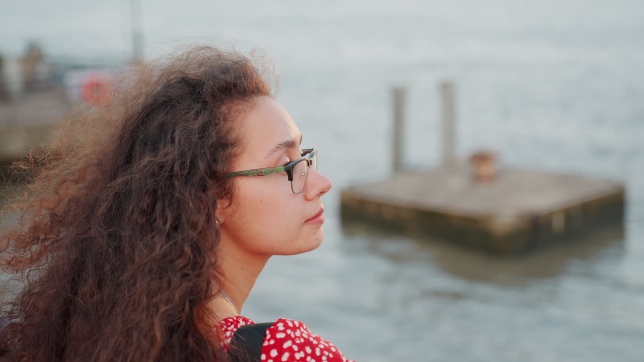 Young woman facing sunlit water, artist on pier with flowing curly hair and glasses, golden reflections on river, distant dock and blurred figures, warm sunset glow and hopeful ambience