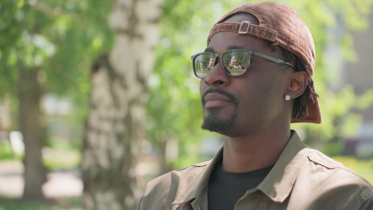 Close up of man adjusting sunglasses while wearing brown cap and jacket in outdoor park setting with green trees soft daylight and blurred background details