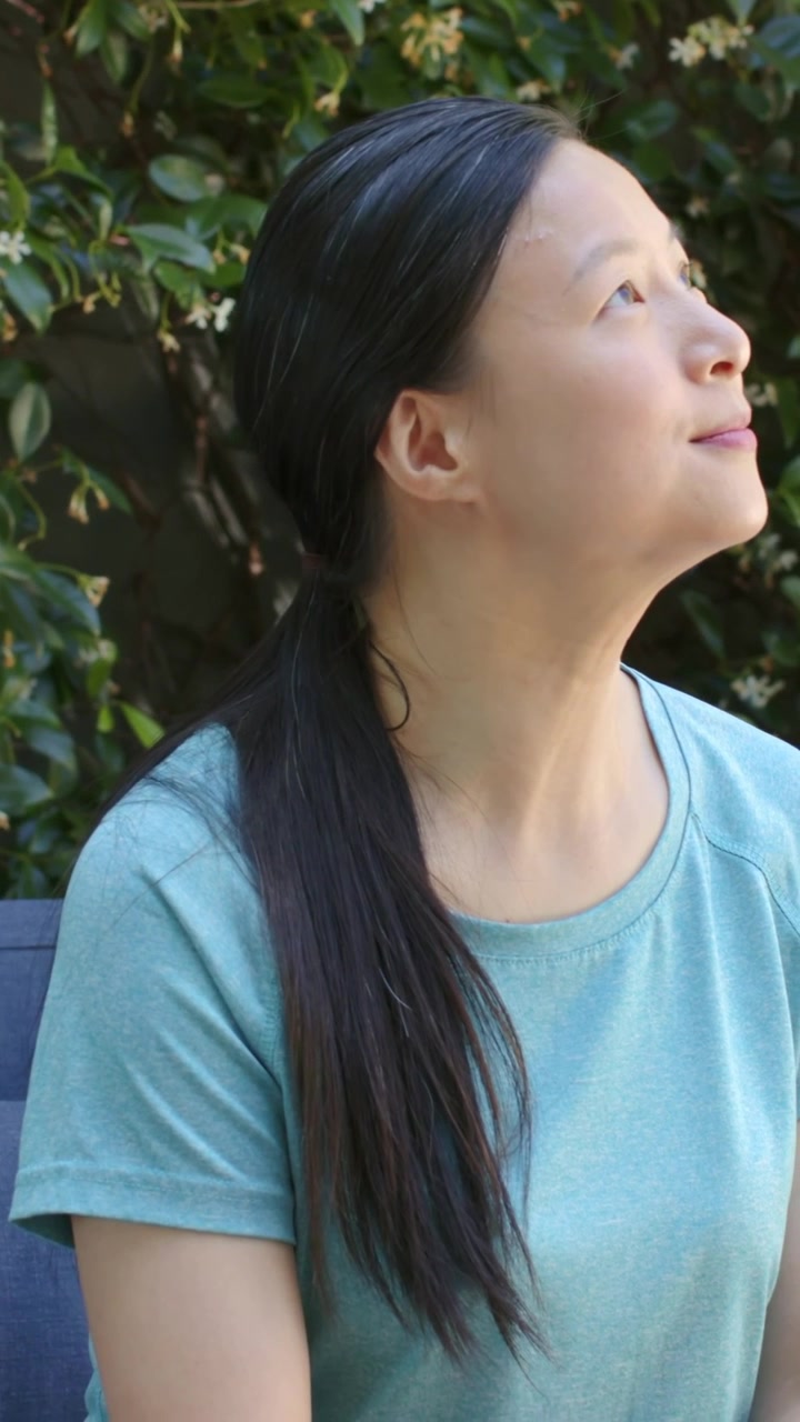縦型動画：首を傾げ、ティール色のTシャツを着た女性が、花咲く生垣のそばの青いベンチに座って上を見上げている。