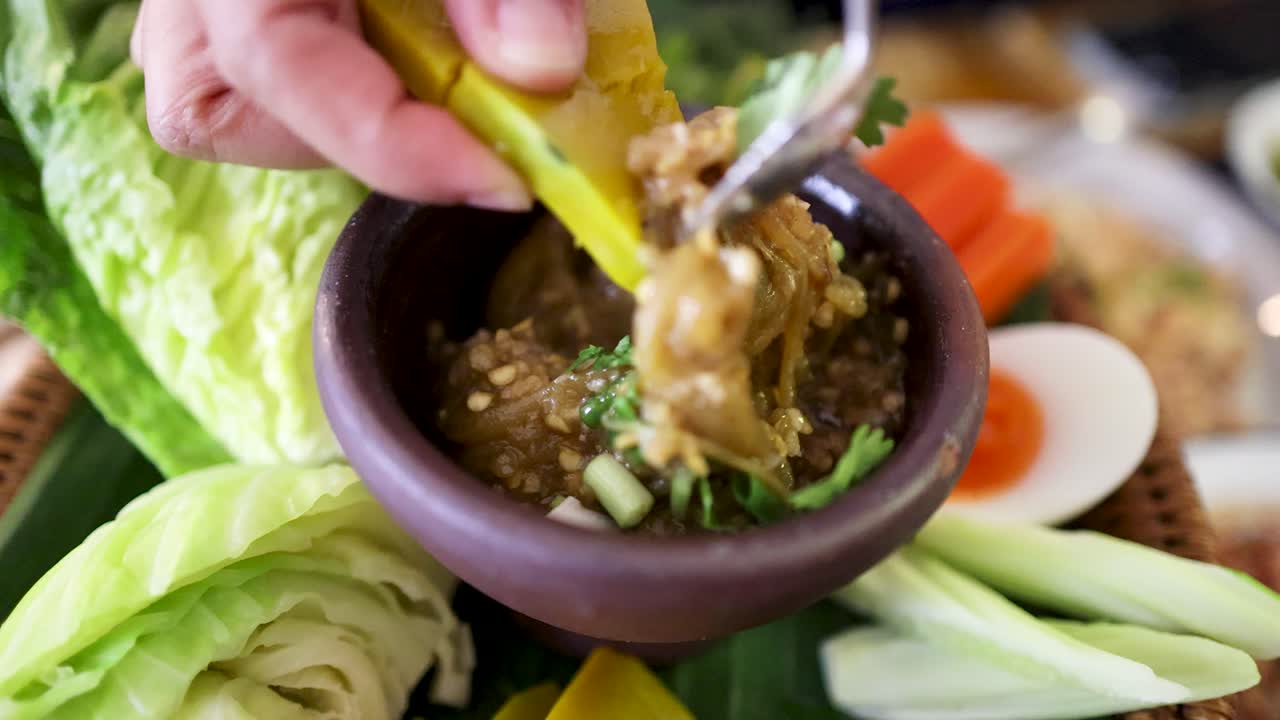 Hand dips boiled pumpkin slice into roasted green chili dip, surrounded by fresh vegetables, cabbage, and boiled egg in natural daylight, close-up view