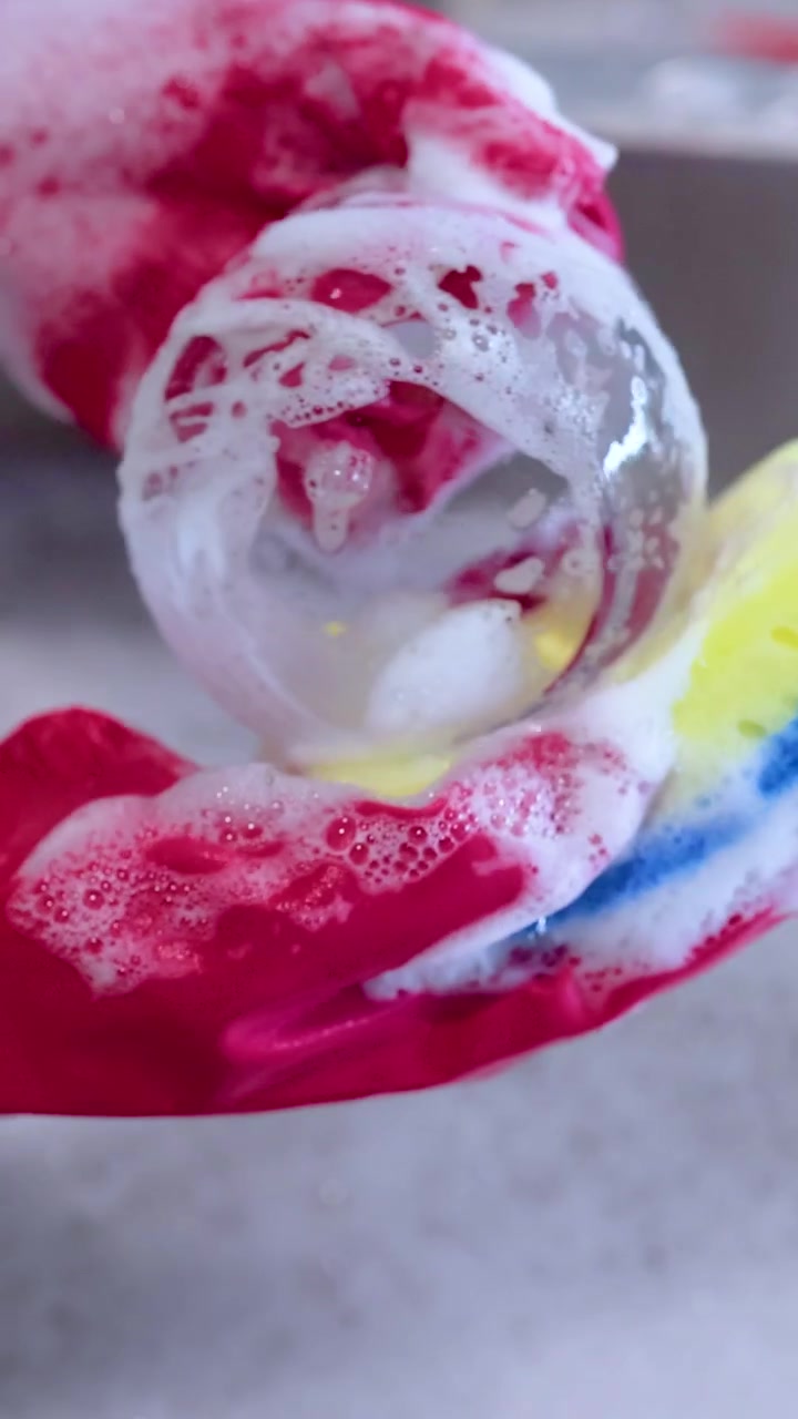Red-gloved hands clean a glass with a soapy sponge in a bright kitchen setting