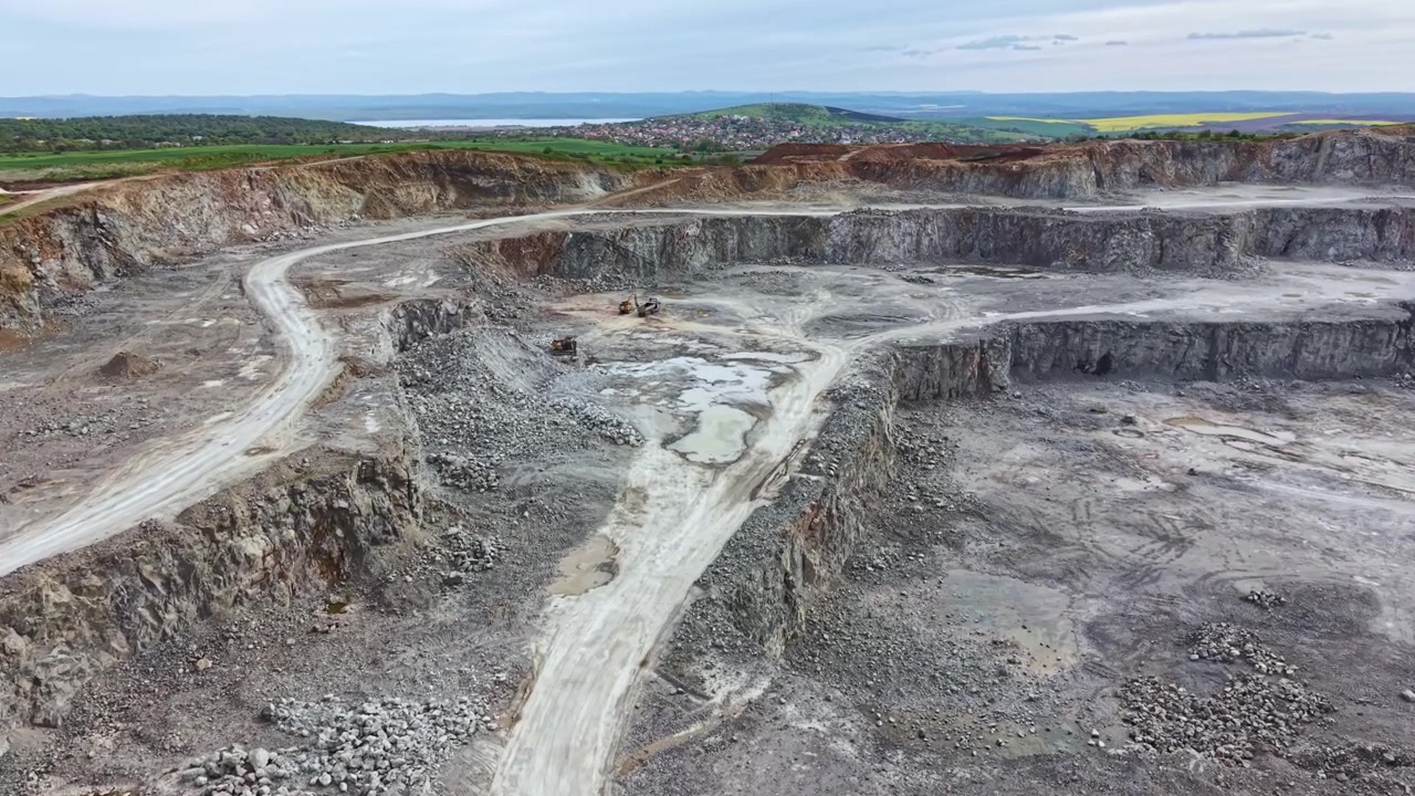 Construction site shows large excavation area with dirt paths and heavy machinery in operation near a town in the countryside during daytime hours