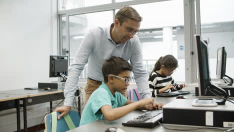 focused schoolboy sitting and typing on keyboard while male teacher standing and looking at student work