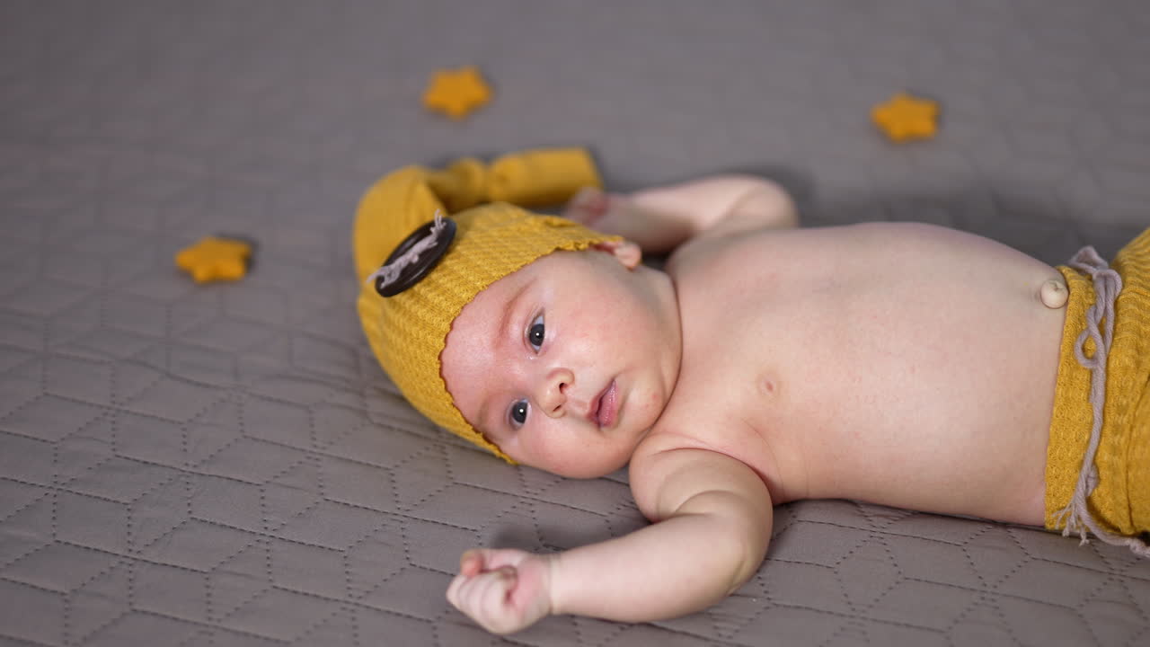 Sweet baby lying on the bed and moving his little fists randomly. Newborn wearing yellow costume and surrounded by toy stars close up.