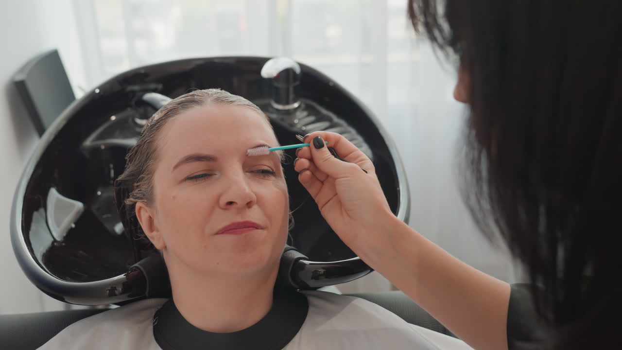 woman reclining at salon shampoo sink, stylist applying eyebrow tint and foam to hairline, client wearing protective cape, relaxed expression, bright natural light, closeup beauty treatment and self
