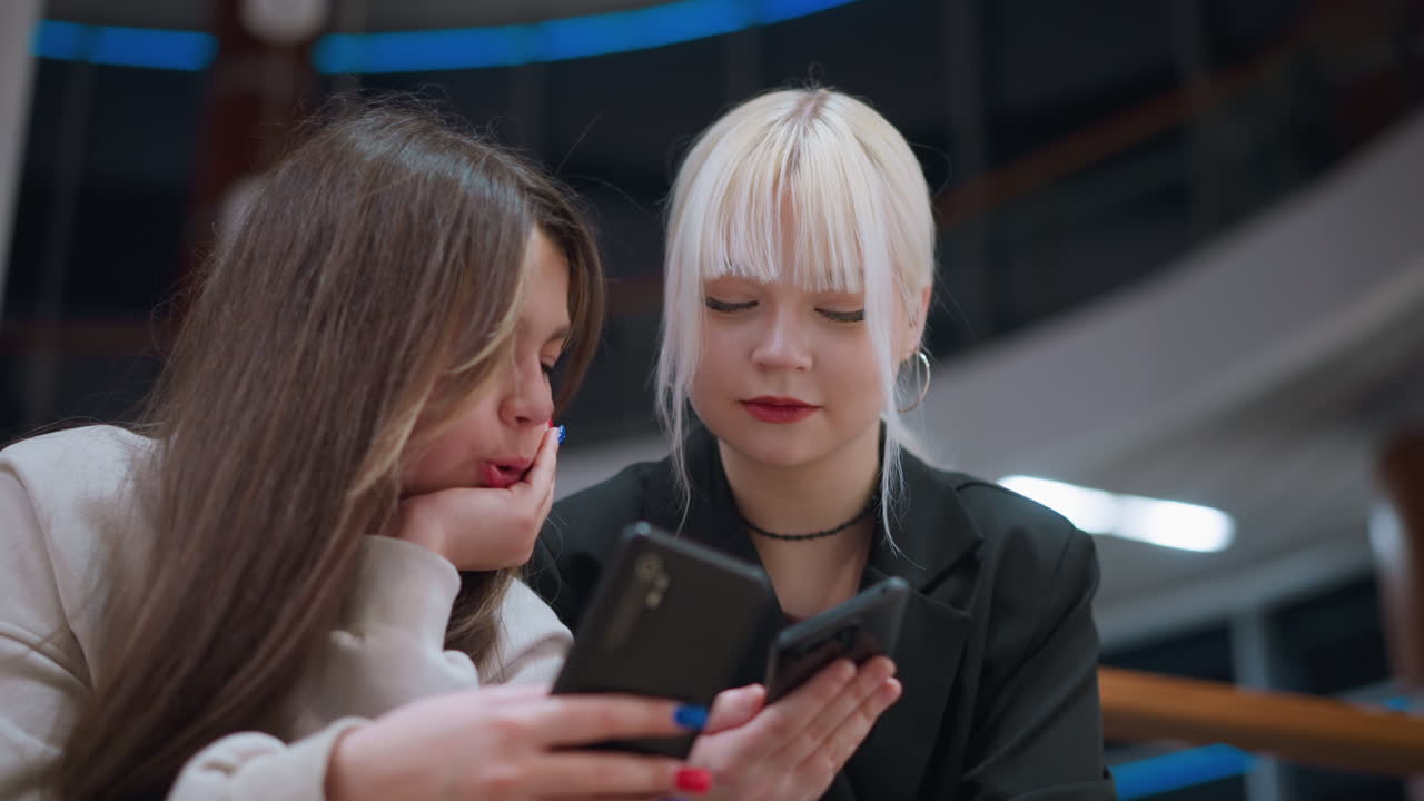 Two teen girls sitting indoors using phones together with focused expressions showing modern lifestyle, digital culture, youth communication, technology dependence and social media engagement