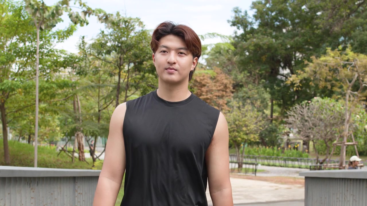 晴れた日、引き締まった体型のアジア人男性が緑豊かな都市公園をジョギングしている。