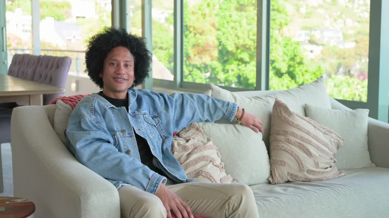 Uomo afroamericano di mezza età seduto sul divano vicino alle finestre che sorride e ride mentre ascolta un dialogo fuori campo