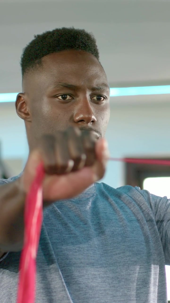 Vertical video: Wearing blue tee, fit man gripping red band, performing pulls in gym for strength