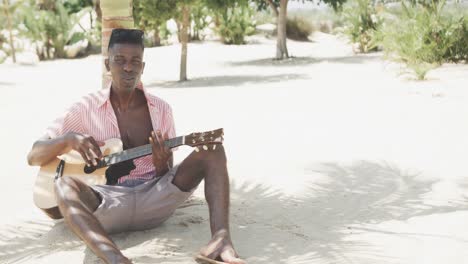 Feliz-Hombre-Afroamericano-Sentado-Bajo-Un-árbol-Tocando-La-Guitarra-Y-Cantando-En-La-Playa-Soleada,-Cámara-Lenta