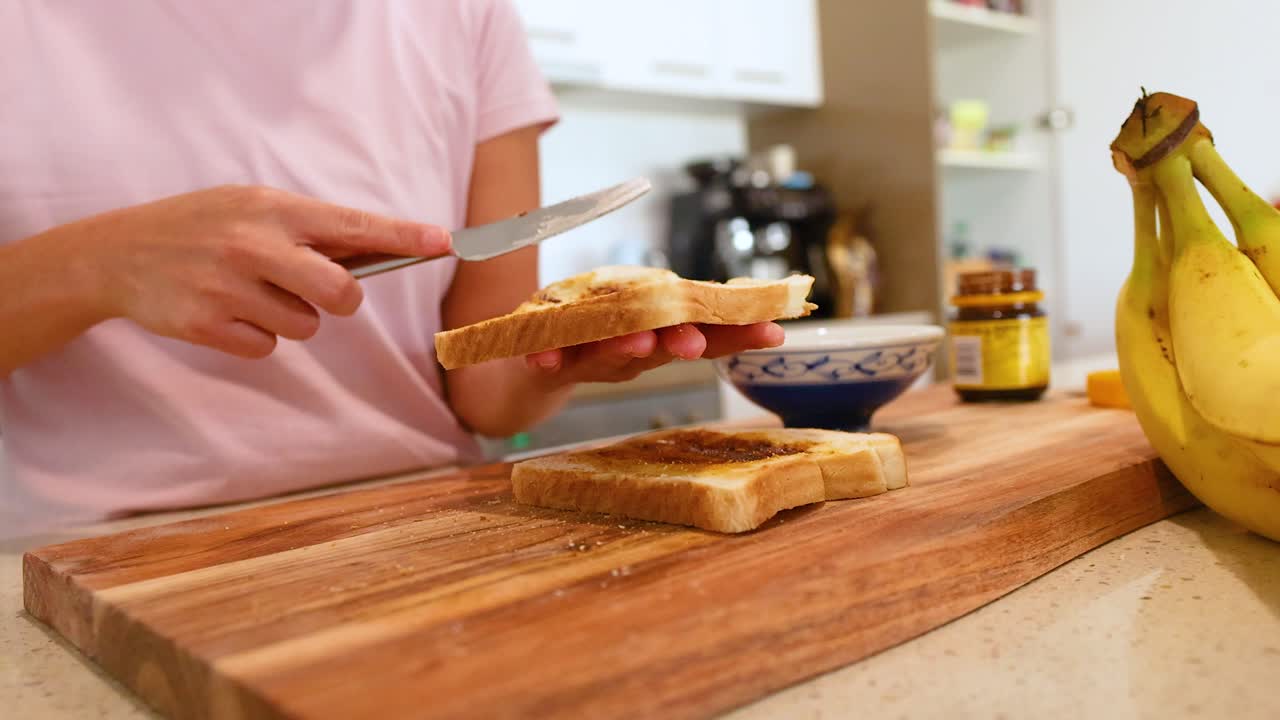 A person spreads Vegemite on buttered toast in a well-lit kitchen, with bananas and a jar nearby