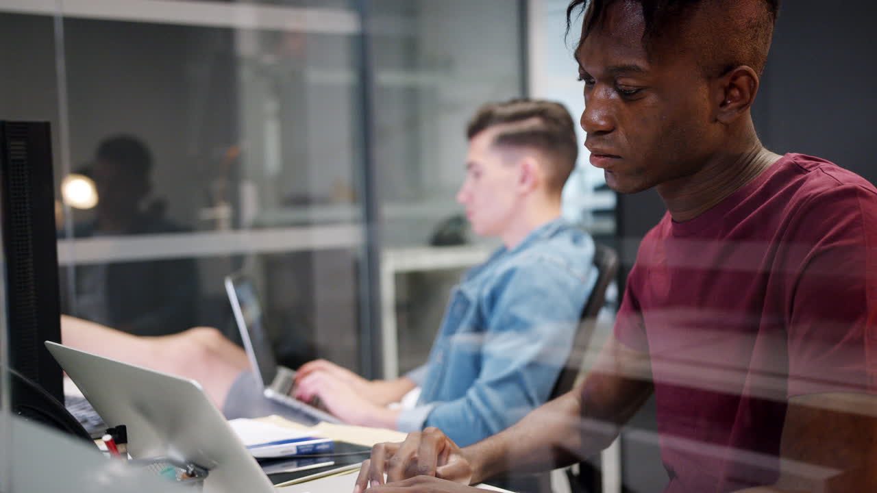 Side view of two young men working at computers sitting in a glass office cubicle, close up, seen through glass
