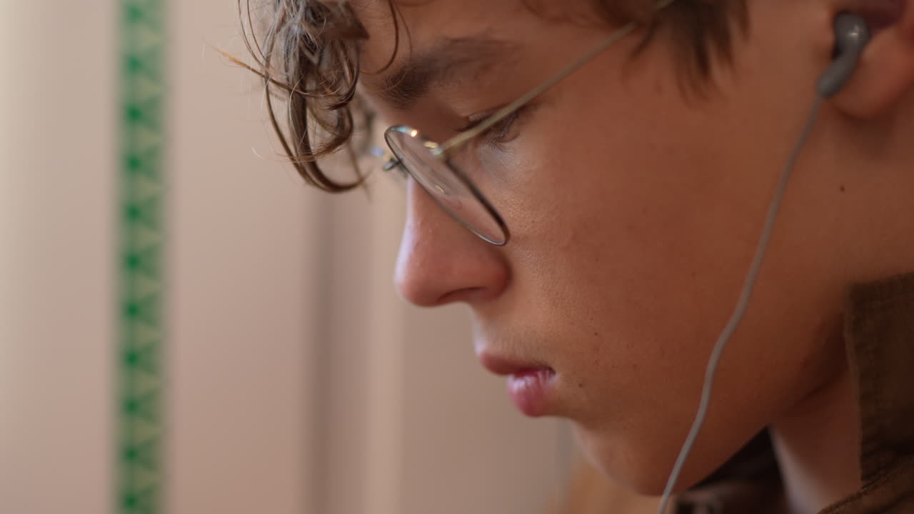 Young male student with glasses listening to music, focused on study in a cozy indoor environment