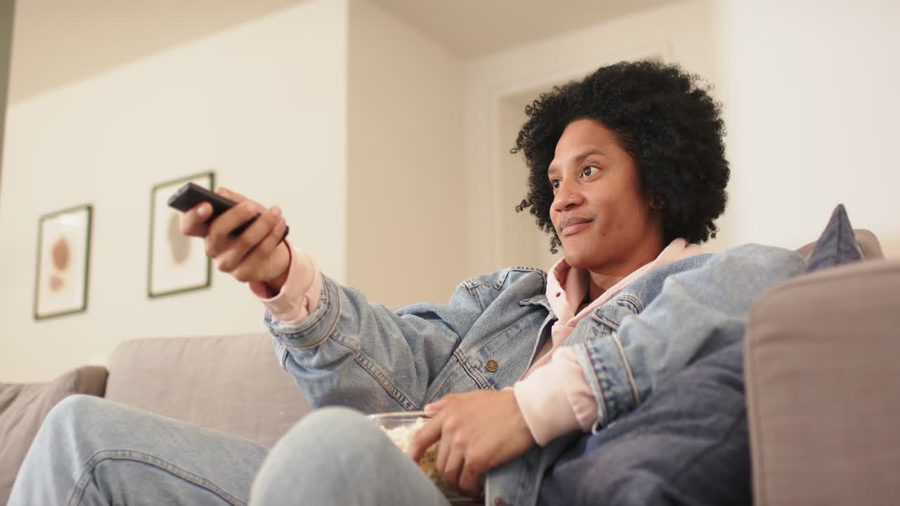 Uomo afroamericano che tiene in mano un telecomando e dei popcorn in salotto, che ride dopo un momento divertente in TV