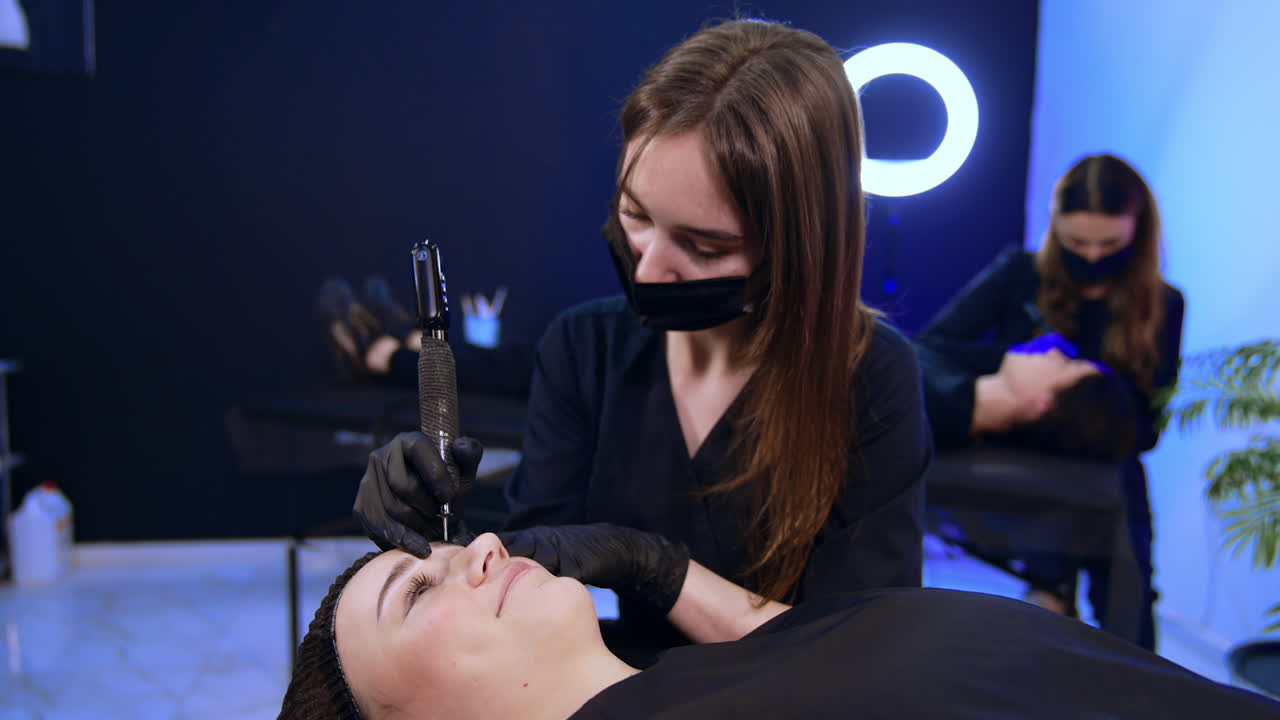 Spacious room in a beauty salon where two beauty professionals work with clients. Beautician perform procedure of permanent eyebrow make up.