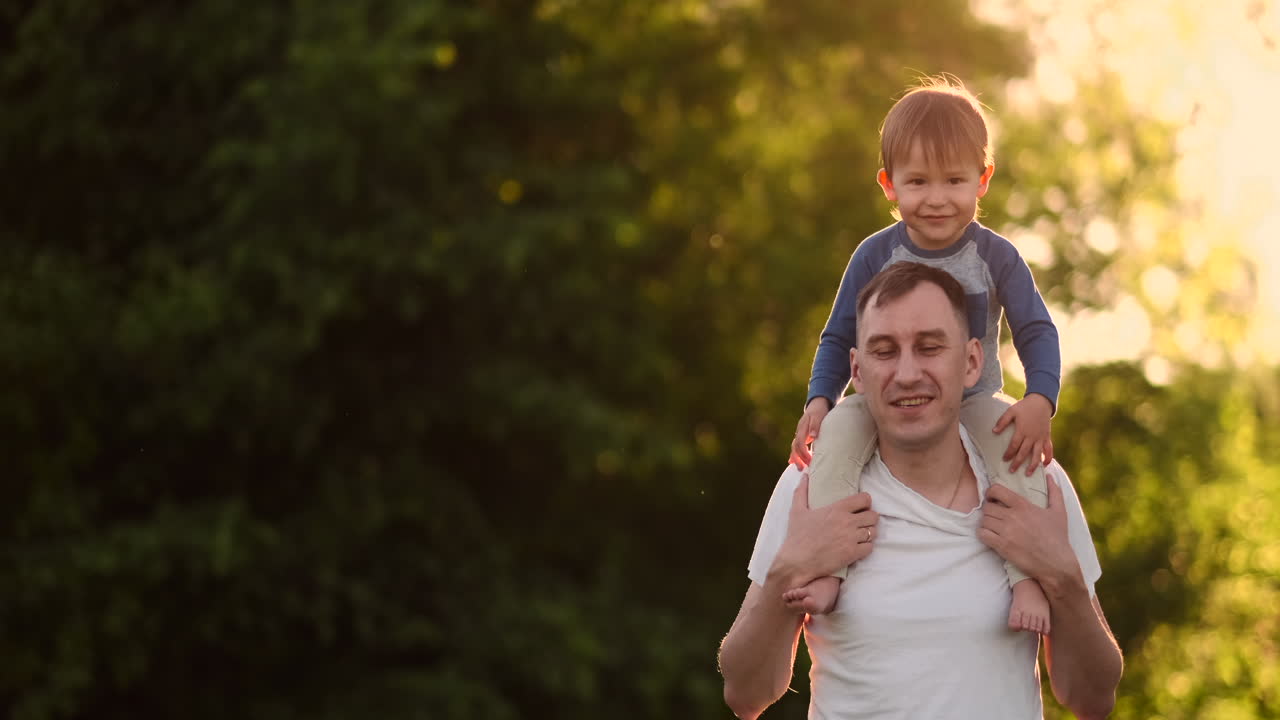 Padre amorevole sorride camminando con il bambino seduto sulle spalle al tramonto su un prato in estate al rallentatore