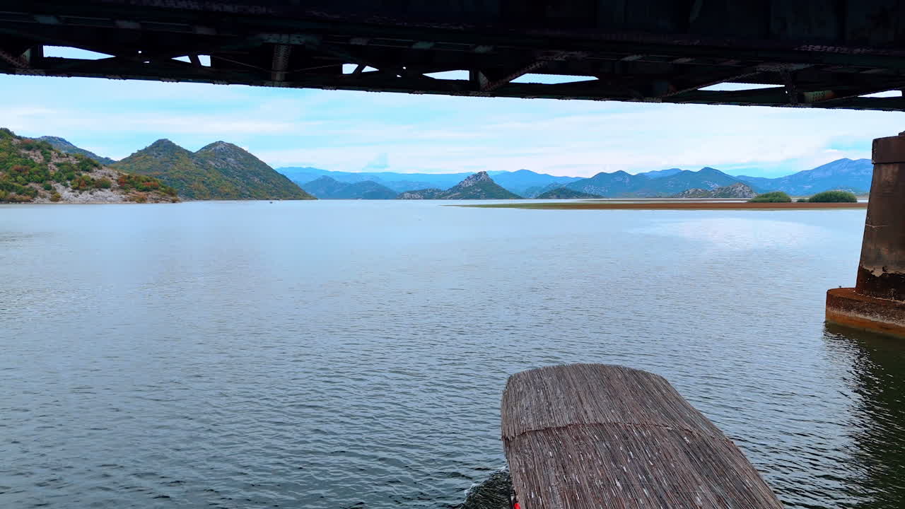 Ulcinj, Montenegro, 14 August 2025: Boat Approaching Old Bridge. A red boat moves toward an old rustic bridge across a quiet lake